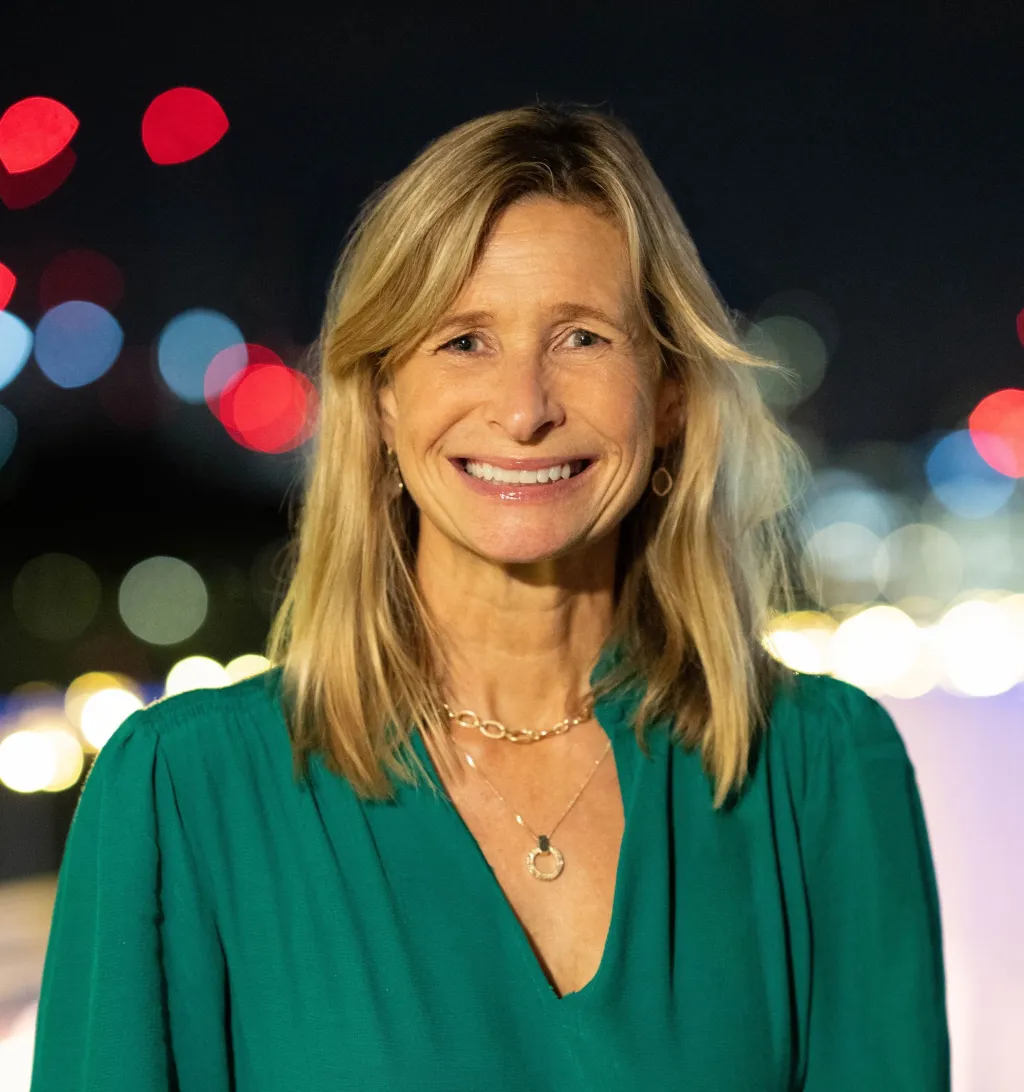 Woman with shoulder-length blonde hair wearing a green blouse and pendant necklace, smiling at the camera against a blurred bokeh background of city lights at night.
