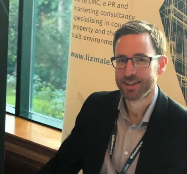 A man wearing glasses and a dark blazer over a light blue shirt smiles at the camera while standing in front a yellow banner about LMC, a PR and marketing consultancy specializing in construction and the built environment.