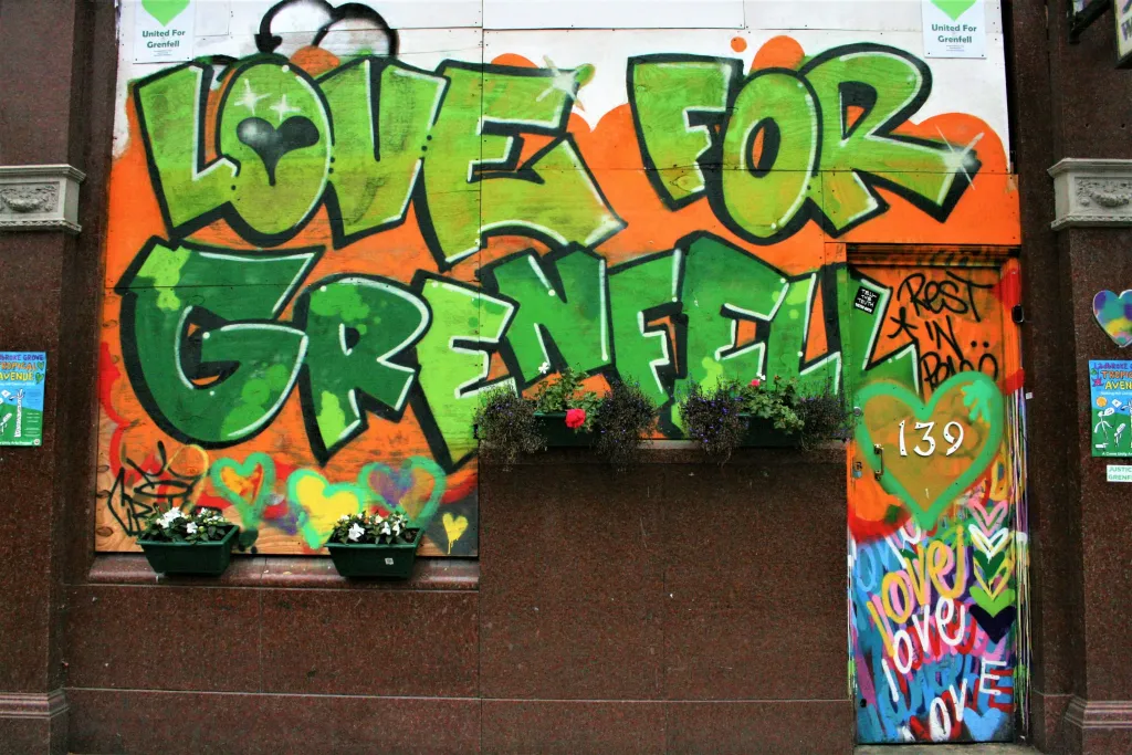 A brick building wall displays vibrant green and orange graffiti reading "LOVE FOR GRENFELL" with decorative flowers in planters beneath it and additional colorful street art visible on the right side.