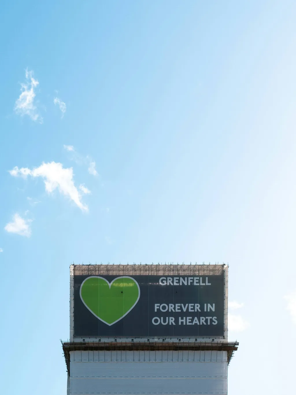 A large billboard mounted on a white tower against a blue sky displays a green heart with 