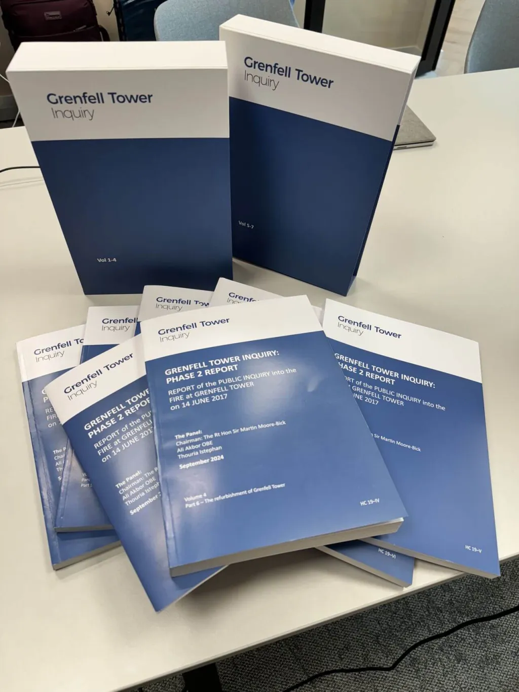 Multiple copies of the Grenfell Tower Inquiry Final Report displayed on a white table, including two large white and navy boxed sets and several individual blue-covered report documents stacked together.