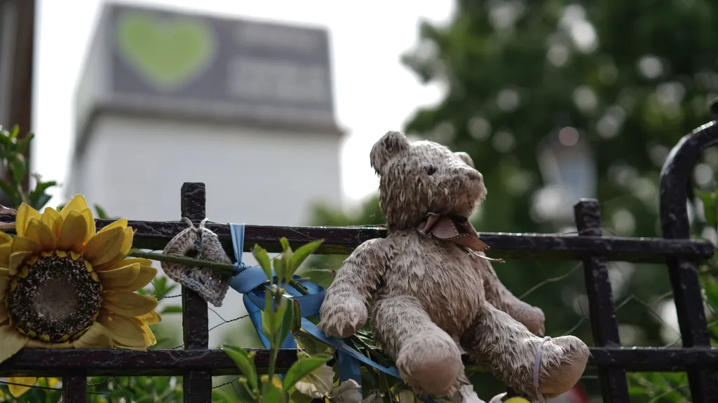 A worn teddy bear sits on a black metal fence next to a yellow sunflower and small blue ribbon, with a building and green foliage blurred in the background.