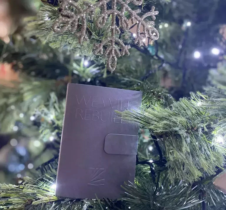 A purple leather wallet or card holder with embossed text sits nestled among evergreen branches and white fairy lights on a decorated Christmas tree.