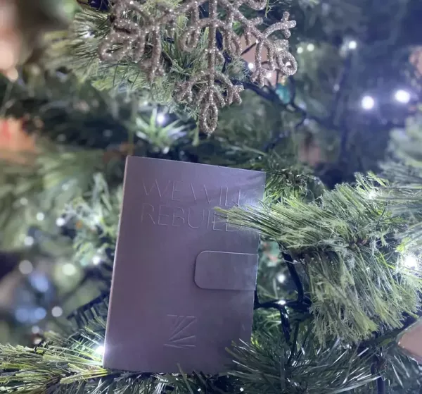 A purple leather wallet or card holder with embossed text sits nestled among evergreen branches and white fairy lights on a decorated Christmas tree.