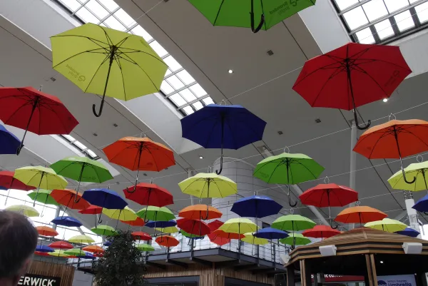 Colorful umbrellas in red, yellow, green, and blue hang from the ceiling of a modern indoor shopping center, creating a vibrant artistic installation beneath skylights.
