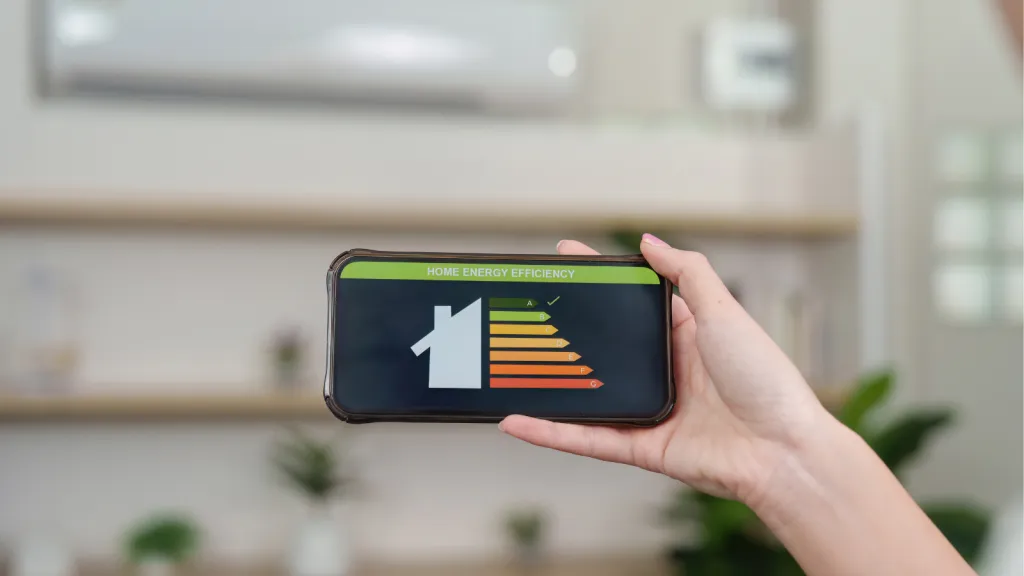A hand holds a smartphone displaying a white house icon with a colorful bar chart in yellow, orange, and red, symbolizing energy efficiency or home energy upgrades against a blurred kitchen background.