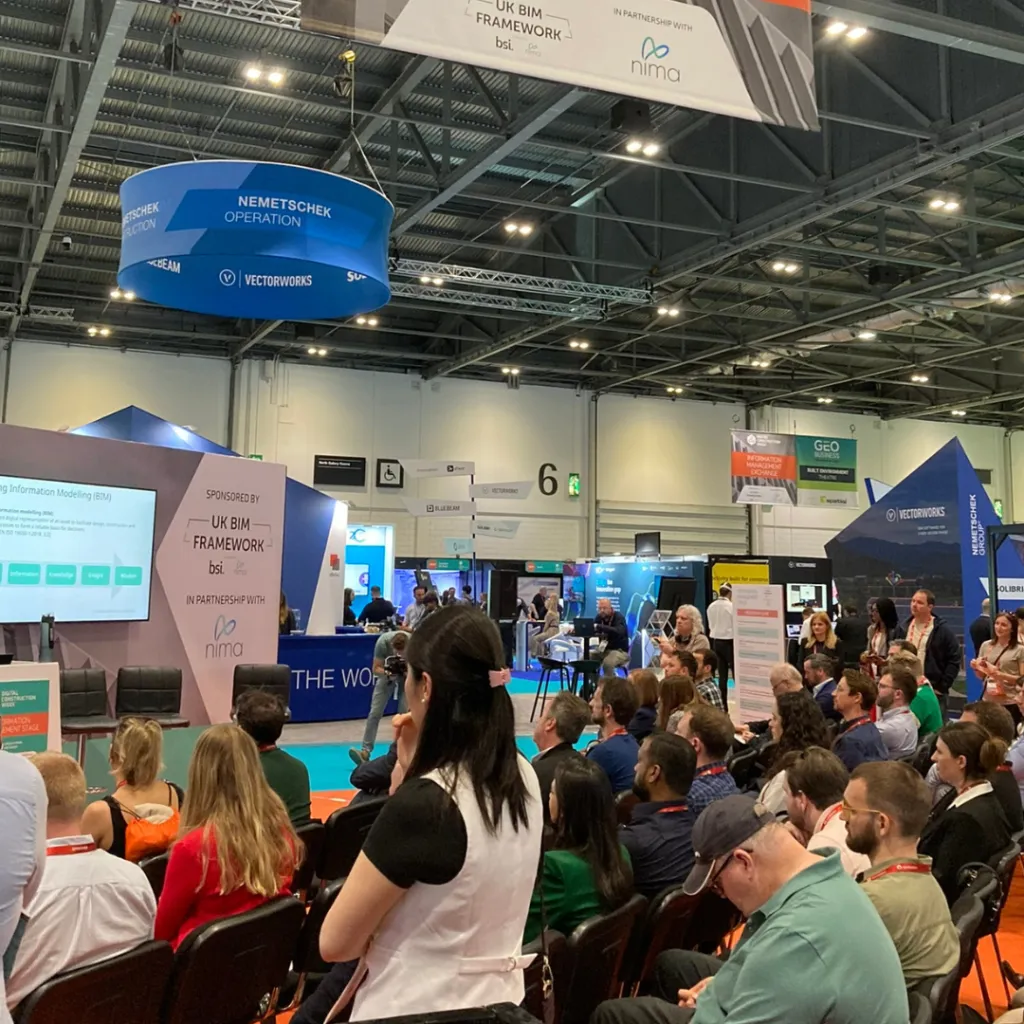 A large indoor conference or trade show with hundreds of attendees gathered in a modern convention center hall. Blue exhibition booths and signage are visible throughout the crowded floor, with overhead banners and bright ceiling lights illuminating the space.