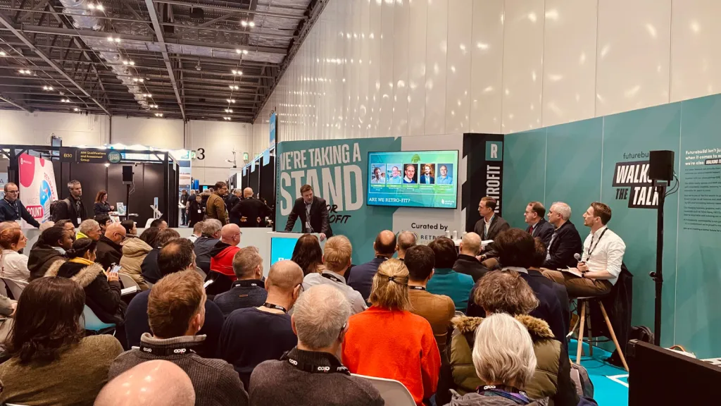 A crowded conference session in a large venue with turquoise-colored branding panels visible. Attendees sit facing a panel of speakers at the front of the room under bright industrial ceiling lights.