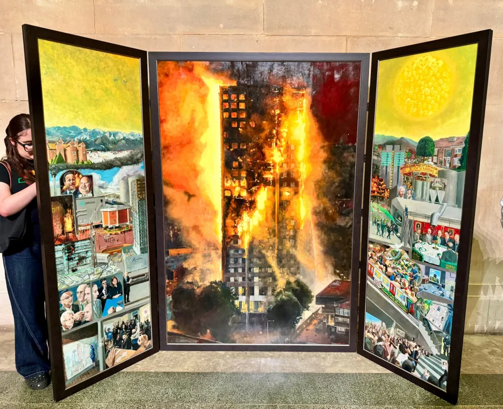 A triptych art installation with black frames displays a burning high-rise building in the center panel, flanked by two panels showing colorful detailed illustrations of urban street scenes with buildings and people. A person in dark clothing stands to the left of the installation.