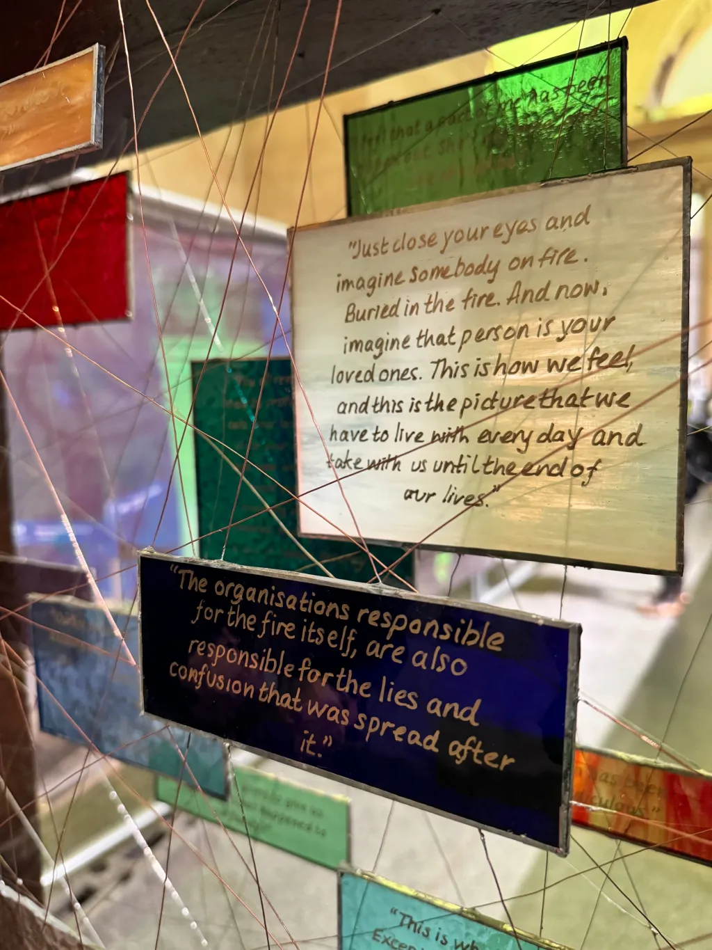 Layered transparent panels with printed text and colored geometric shapes create an artistic installation. The visible text includes reflective passages about loss and organizational responsibility related to the Grenfell tragedy, with strings connecting the panels in a web-like pattern that symbolizes interconnection and complexity.
