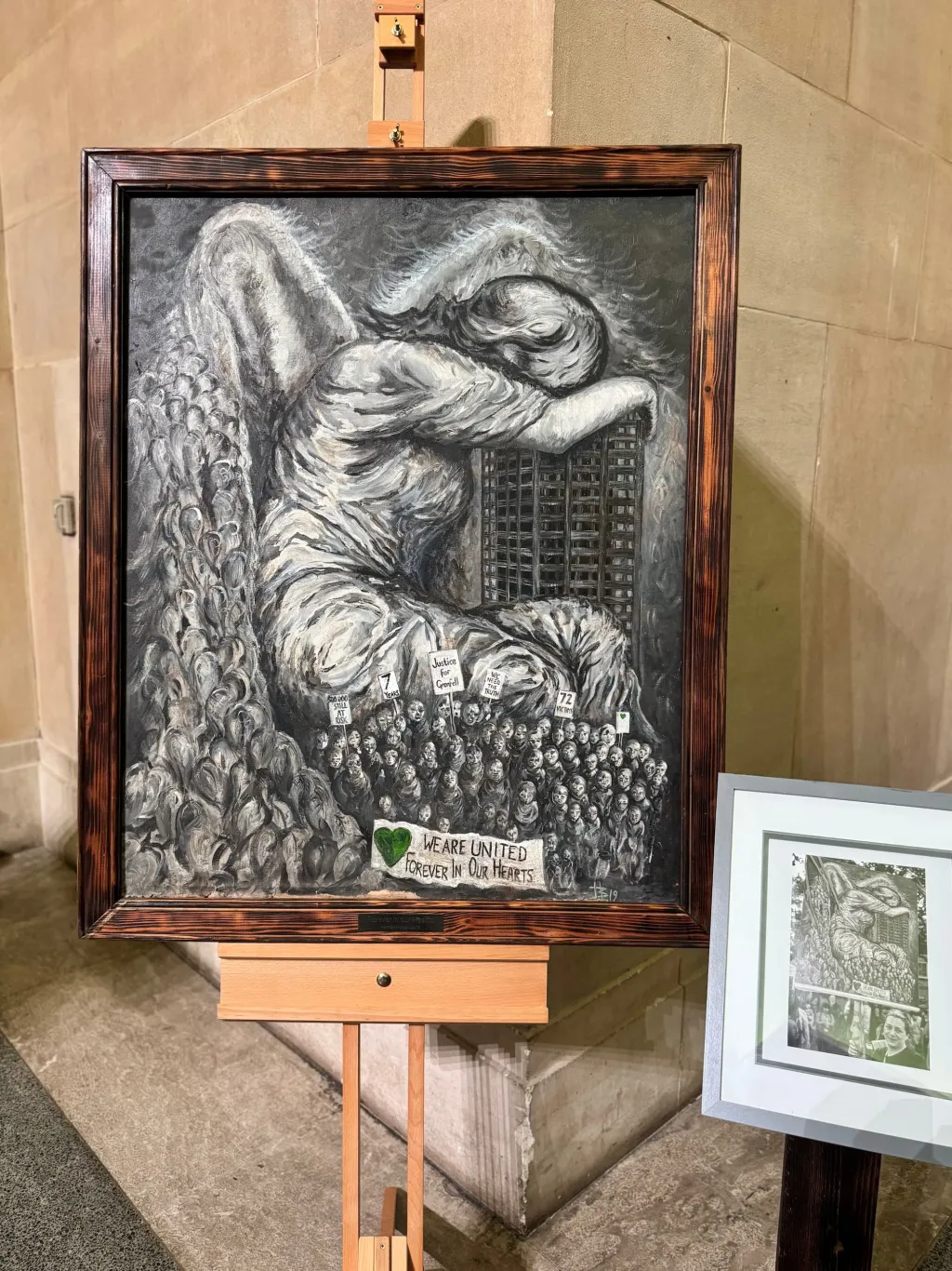 A detailed black and white etching depicts an angel with large wings looking down at a vast crowd of people below, mounted in a wooden frame on a wooden easel in what appears to be a gallery or exhibition space.