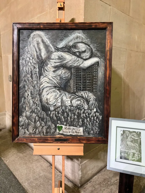 A detailed black and white etching depicts an angel with large wings looking down at a vast crowd of people below, mounted in a wooden frame on a wooden easel in what appears to be a gallery or exhibition space.