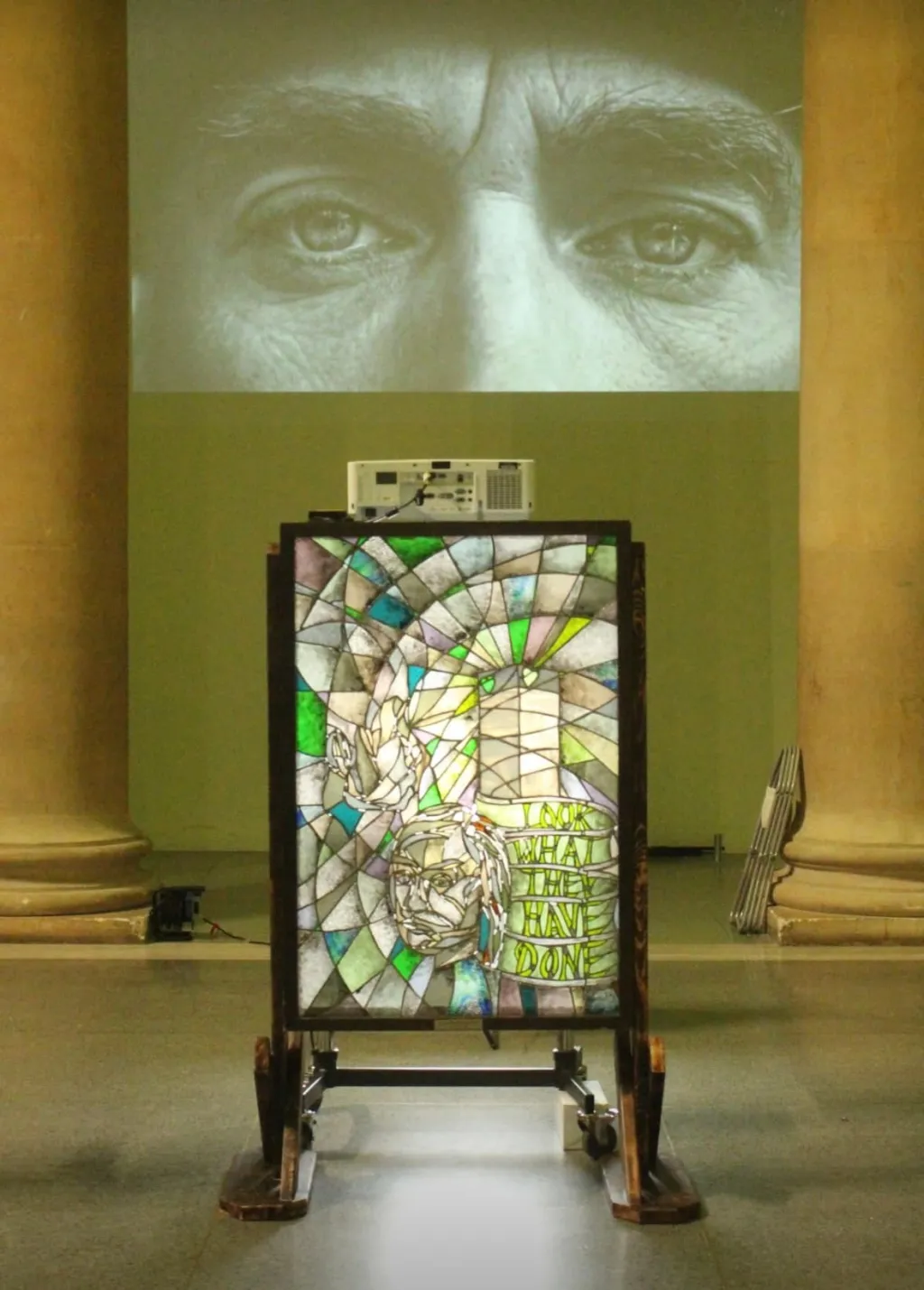 A gallery installation featuring a large stained glass window panel with geometric patterns in greens and earth tones, mounted on a wheeled stand beneath a projected close-up of human eyes and a green wall. The artwork appears to be part of an exhibition exploring perspectives on the Grenfell Tower tragedy.