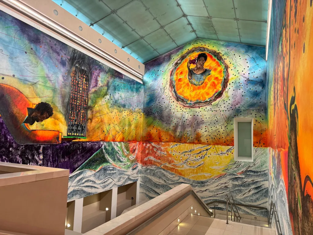 A vibrant mural covering the walls and ceiling of an indoor space, dominated by warm yellows, oranges, and blues, with a large circular sun-like form at the center top and flowing wave patterns at the base, alongside architectural sketches and figures rendered in mixed media style.