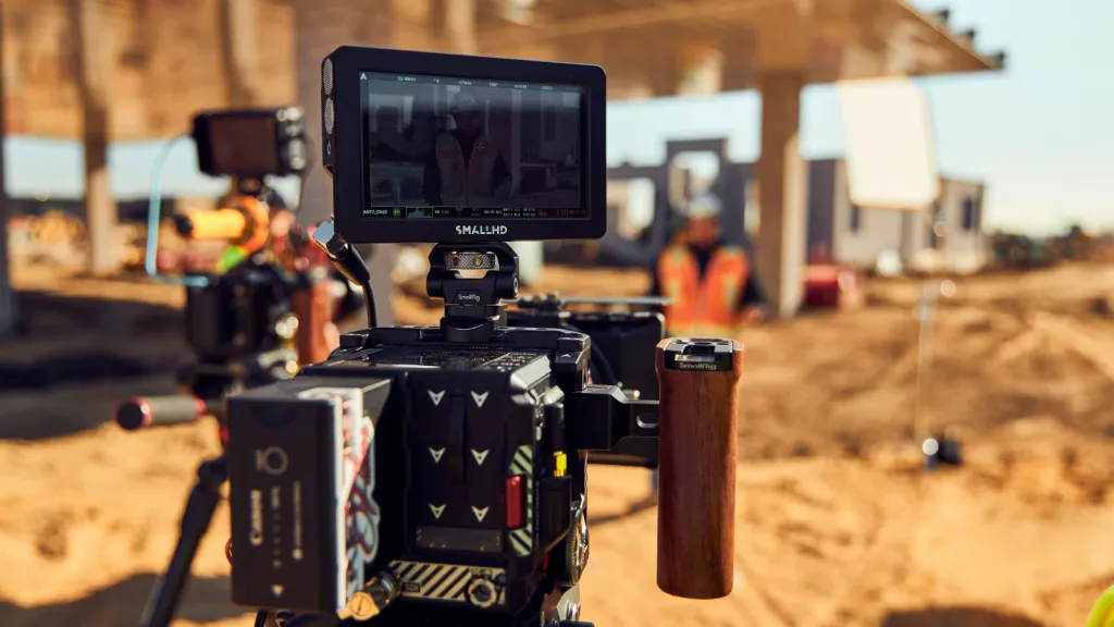Professional video camera rig with external monitor mounted on stabilizer equipment positioned on a construction site, with heavy machinery and buildings visible in the background.