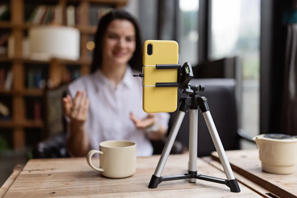 A content creator sits behind a wooden desk with a yellow smartphone mounted on a tripod, gesturing while recording. Two ceramic mugs and a blurred bookshelf are visible in the background of her home studio setup.