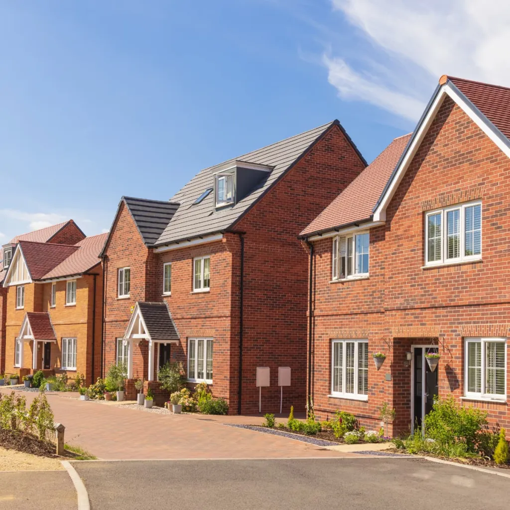 A row of modern brick residential houses with varying heights and roof styles, featuring large windows and well-maintained front gardens along a paved driveway under a clear blue sky.