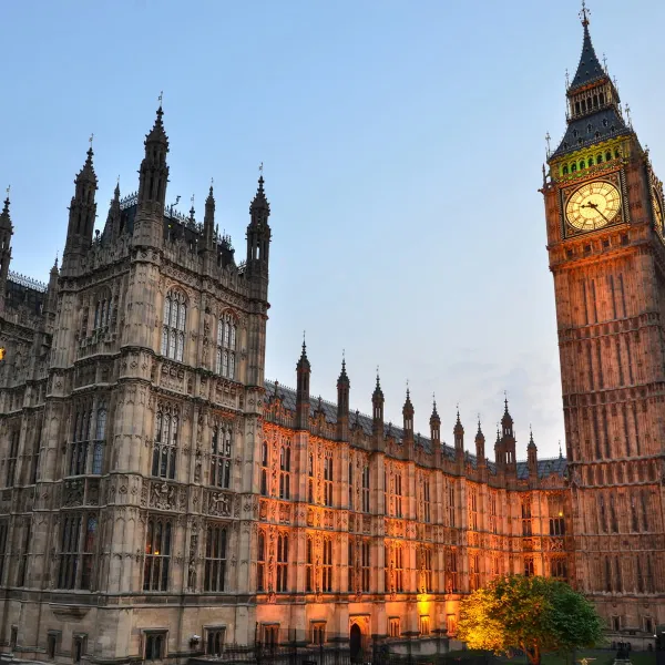The Palace of Westminster at dusk, with Big Ben illuminated on the right and the Gothic Revival architecture of the Houses of Parliament bathed in warm orange light against a blue sky.