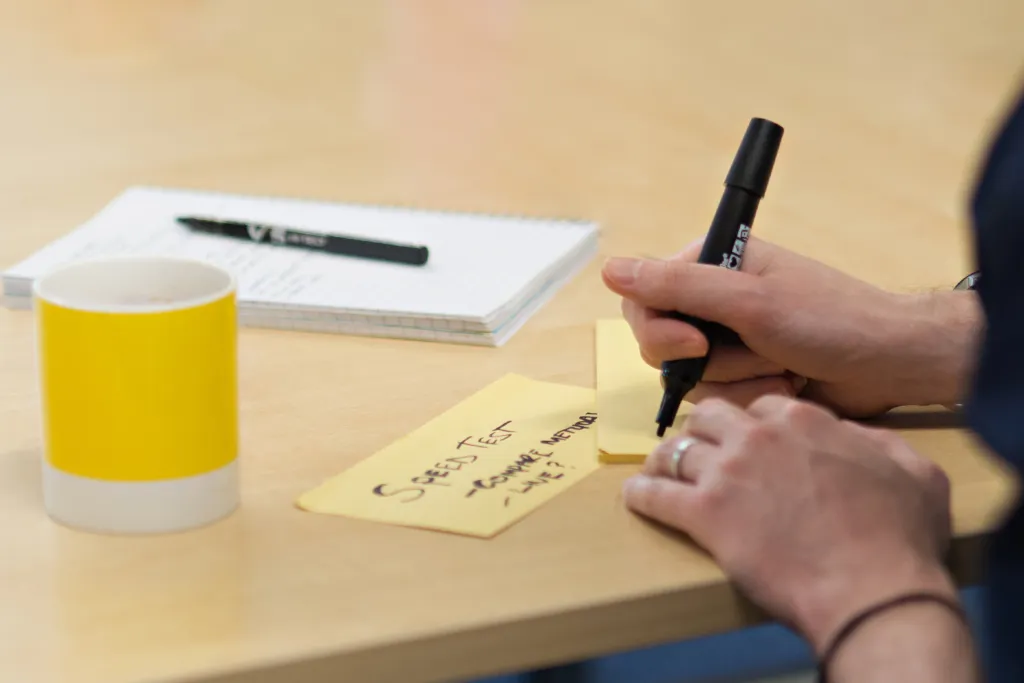 A person writes on a yellow sticky note with a black pen at a wooden desk, with a yellow and white mug and white notepad visible in the background.
