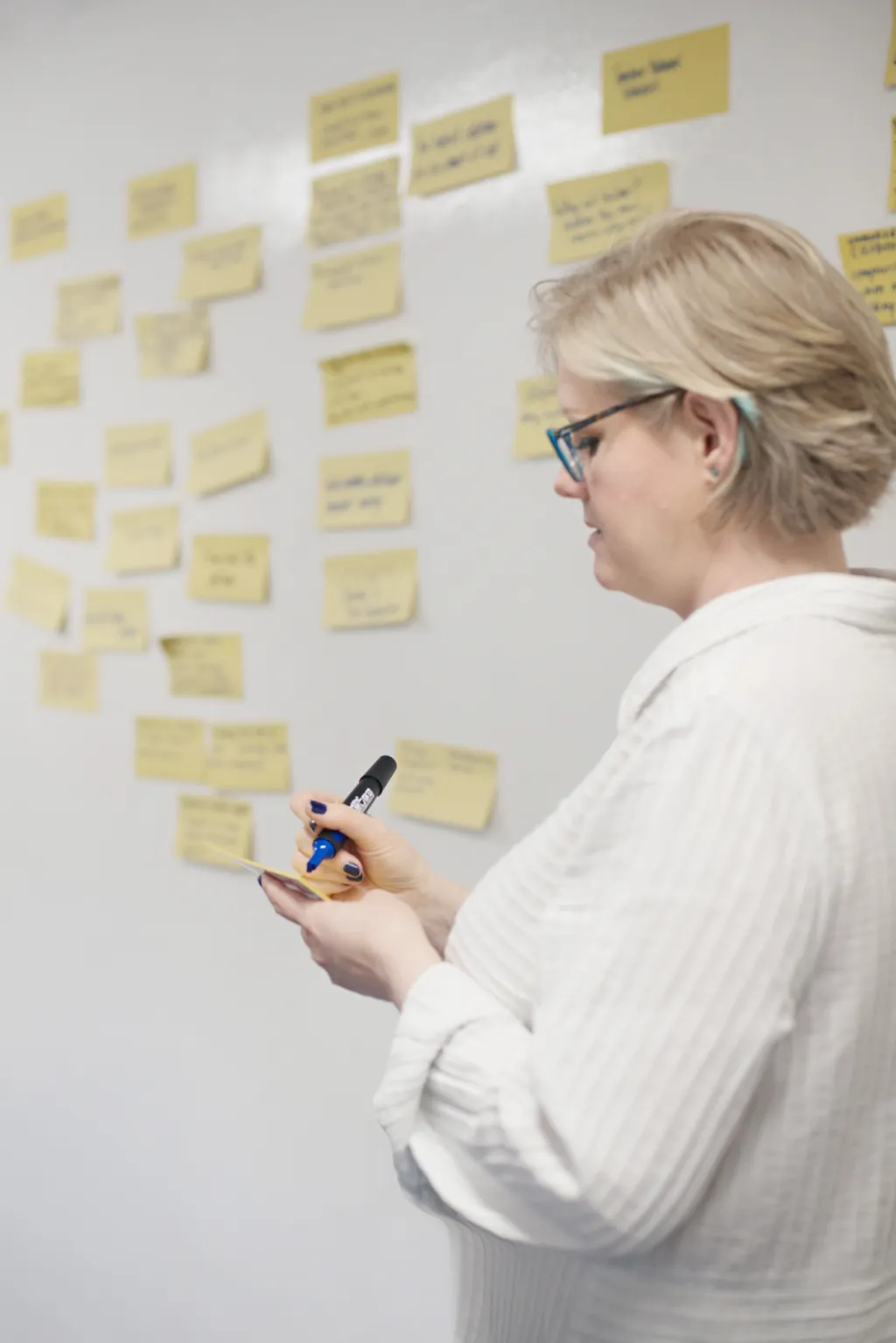 A woman wearing glasses and a white striped shirt holds a pen while reviewing yellow sticky notes arranged on a white wall, appearing to be planning or organizing ideas.