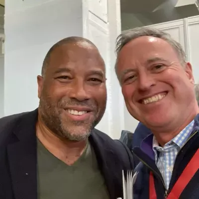 Two men smiling at the camera in an indoor setting, one wearing a dark blazer over a green shirt on the left, and one wearing a blue checkered shirt with a red lanyard on the right.