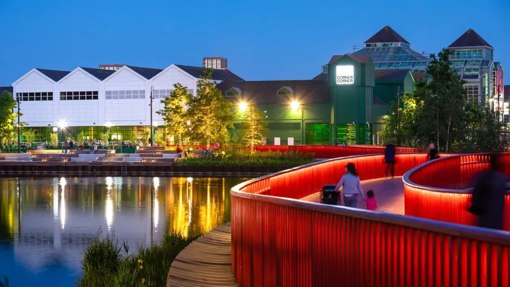 A modern waterfront scene at dusk features illuminated buildings reflected in still water, with a distinctive red curved metal structure in the foreground containing a person, connected by a wooden boardwalk.