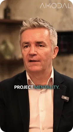 A professional man with gray hair wearing a dark blazer and white shirt speaks against a blurred interior background. Text overlay reads "PROJECT MENTALITY" with the Amodal logo visible at the top.
