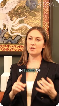 A woman with long reddish-brown hair wearing a black blazer over a white top gestures while speaking, with decorative artwork visible in the background and "IN TERMS OF" text overlaid at the bottom.