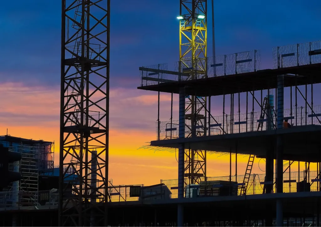 Construction site silhouetted against a dramatic sunset sky, with tall cranes and steel framework of a multi-story building under development illuminated by golden and orange hues.