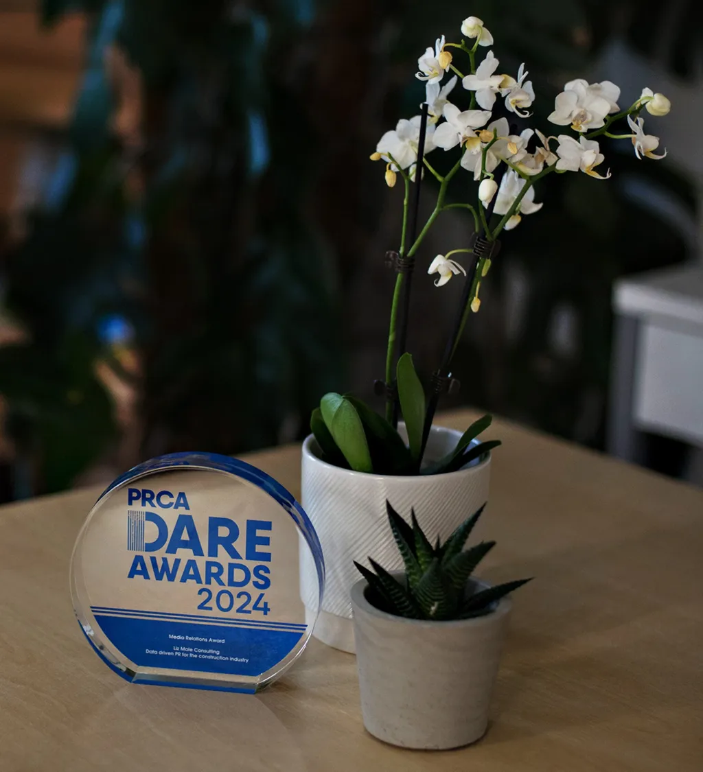 A PRCA Dare Awards 2024 acrylic award sits on a wooden table next to a potted plant with white flowers and dark spiky foliage in white ceramic containers.