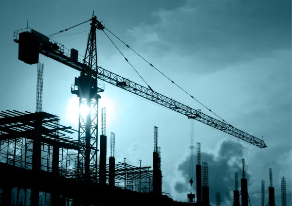 A construction site silhouette against a stormy blue-grey sky features a tall tower crane with its jib extended over steel framework and reinforced concrete pillars. Multiple construction cranes and metal scaffolding are visible in the industrial scene, suggesting an active building project during overcast weather conditions.