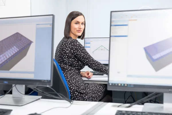 A woman in a dark polka dot jacket sits at a desk with multiple monitors displaying architectural designs and technical drawings of timber structures.