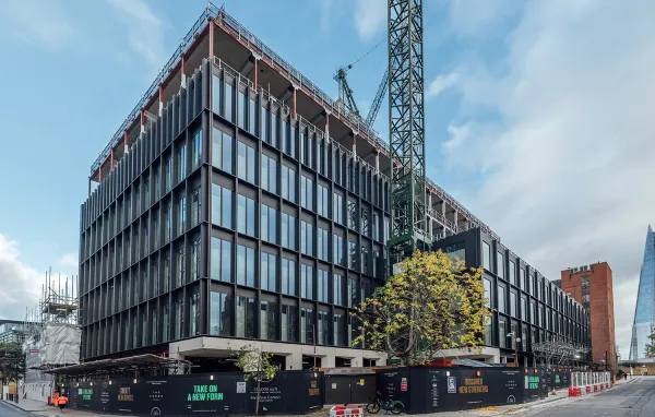 A modern multi-story building with a dark steel frame and large window panels under construction, featuring temporary barriers and scaffolding, with a tall construction crane visible beside it against a cloudy sky.