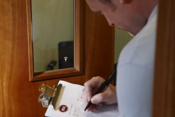 A person in a light blue shirt writes on a clipboard while inspecting a wooden-framed door with a WiFi-enabled smart lock during a fire door safety check.