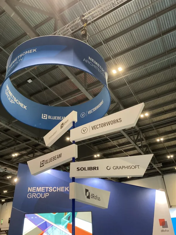 A tall cylindrical blue and white trade show booth display stack featuring company logos including Bluebeam, Vectorworks, Solibri, Graphisoft, and Németschek Group, suspended from the metal ceiling structure of an indoor exhibition hall.