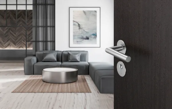 A modern living room with a gray sectional sofa, round metallic coffee table, and light wood flooring, viewed through an open dark door with a contemporary chrome handle and lock visible in the foreground.