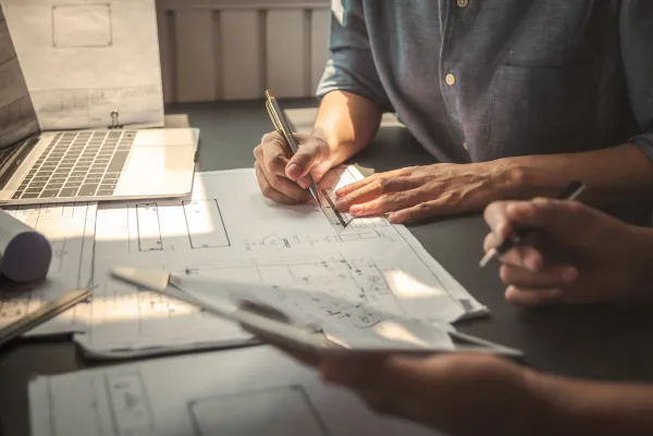 Two people review architectural blueprints and sketches spread across a desk, with one person pointing at details while holding a pen, with a laptop visible in the background.