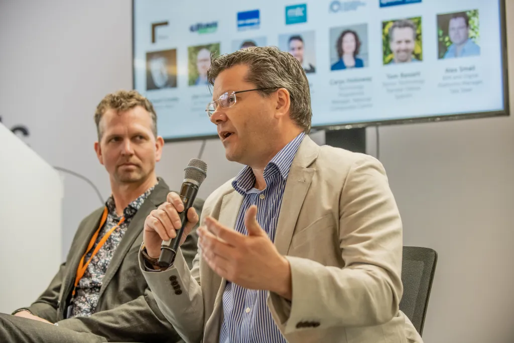 Two men sit on a panel at a professional event, with the man on the right speaking into a microphone while gesturing expressively. Both wear business casual attire, and a screen displaying team member profiles is visible behind them. An orange conference badge is visible on the man on the left.