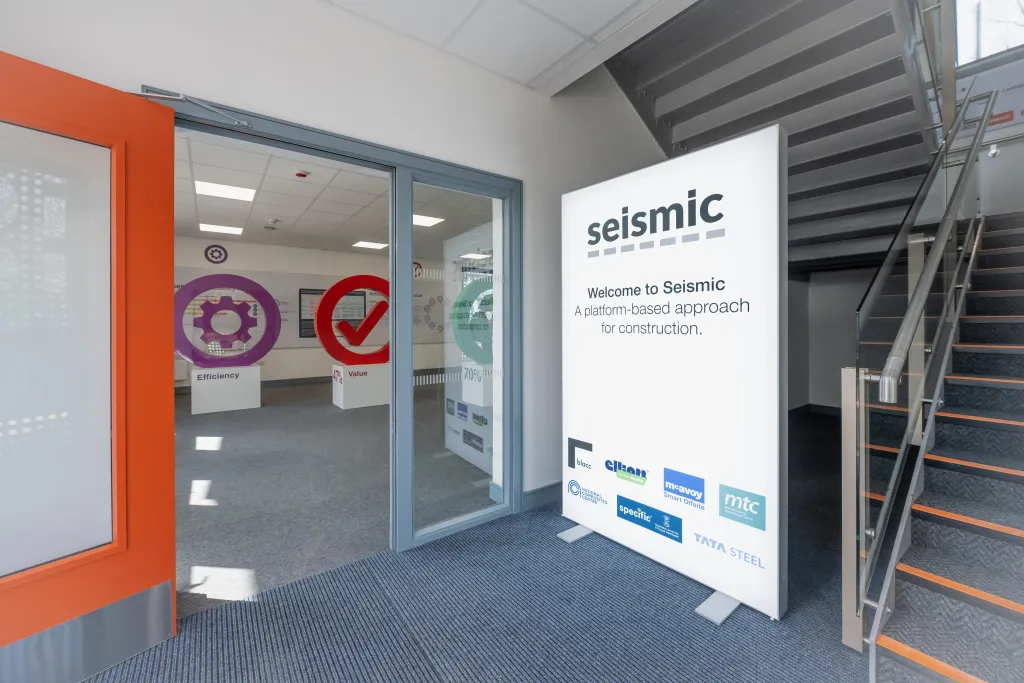 The entrance to Seismic's office features an orange-framed glass door with two circular logos, alongside a white welcome banner listing partner companies including GitHub, AWS, and Stripe. A metal staircase is visible to the right of the entrance.