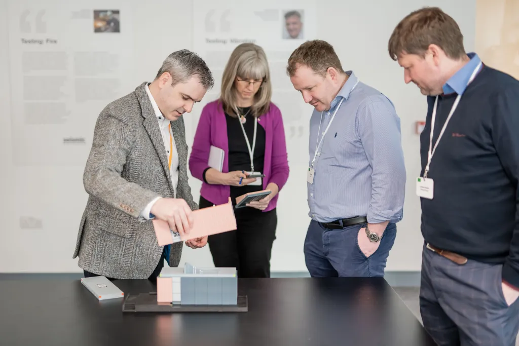 Four professionals wearing ID badges gather around a table examining a tablet device during what appears to be a business meeting or conference presentation.