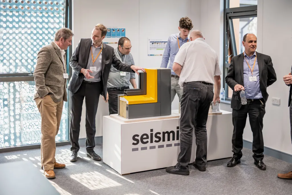 Several business professionals gather around a yellow and dark blue display booth branded with "seismi" at what appears to be a corporate event or trade show. The attendees are dressed in business casual attire and engaged in conversation near the booth. Large windows with a geometric pattern are visible in the background of the modern indoor venue.