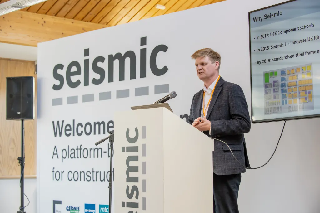 A man in a dark blazer presents at a conference, holding a microphone while standing beside a large white banner displaying "seismic" and text about a platform for construction. A presentation screen is visible in the background to the right, with wooden ceiling panels and a speaker visible in the upper left.
