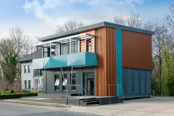 A modern two-story demonstration building with corrugated metal siding in copper and blue tones, featuring large windows, a turquoise canopy entrance, and a metal staircase, set against bare trees and clear sky.