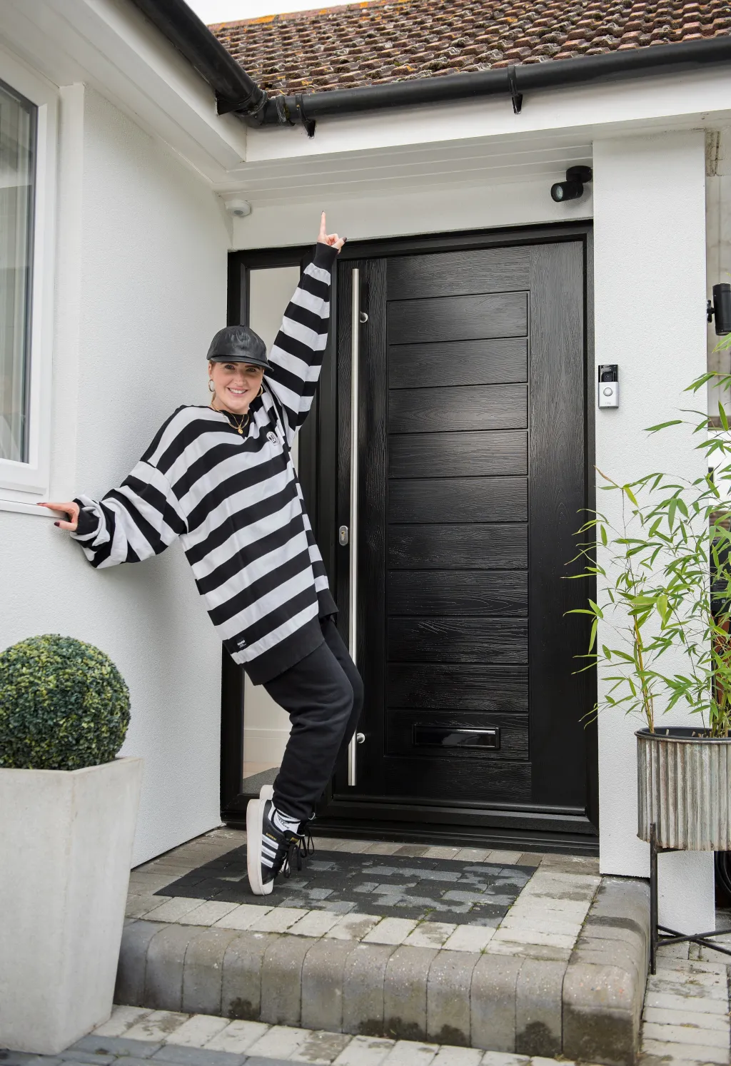 A person in a black and white striped outfit and hat playfully leans against a modern black front door with horizontal paneling on a white-walled porch, flanked by potted plants and decorative bushes.