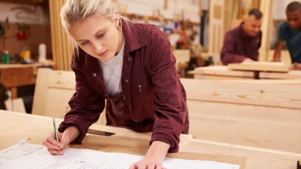 A woman with blonde hair wearing a maroon jacket marks measurements on a wooden board in a workshop while other workers are visible in the background.