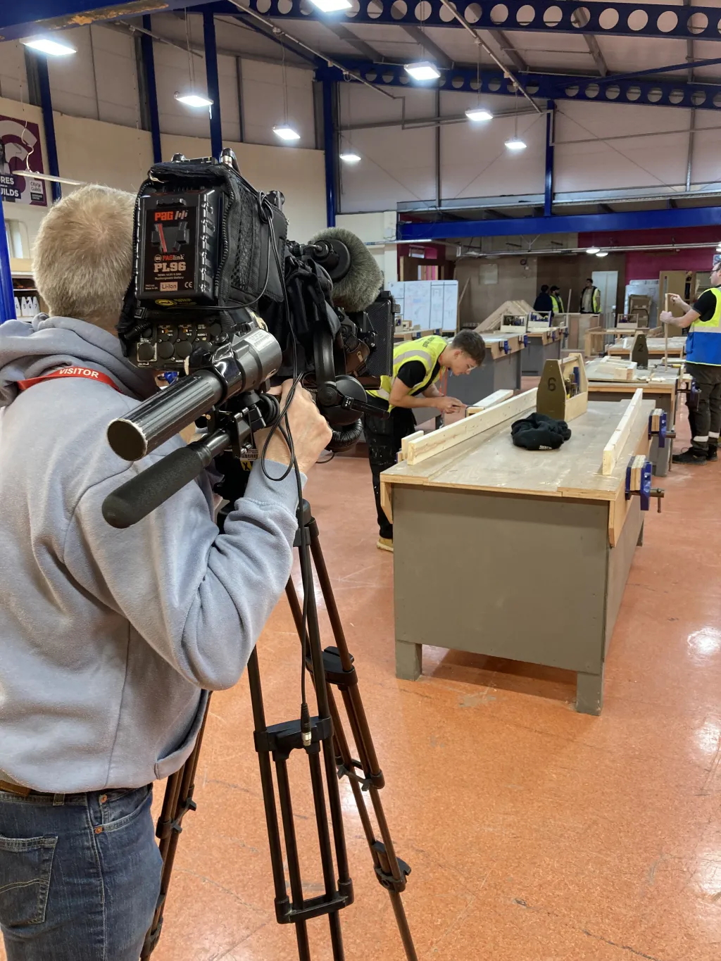 A cinematographer operating a professional video camera on a tripod films inside a construction or workshop facility, with tables, equipment, and workers visible in the background under industrial lighting.