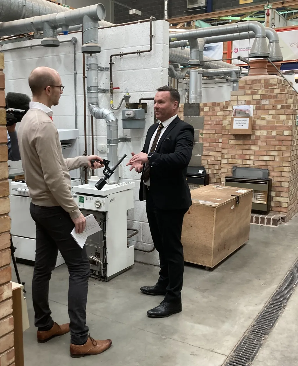 Two men in business attire stand in an industrial warehouse setting, examining HVAC equipment. The man on the left holds documentation while the man on the right gestures toward a large white heating or cooling unit mounted on a brick wall.