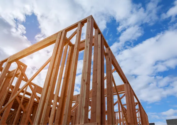 Wooden timber frame structure under construction against a blue sky with white clouds, showing vertical and diagonal support beams typical of residential building frameworks.
