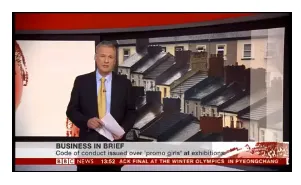 A news presenter in a dark suit and yellow tie stands in a television studio next to an animated graphic of modern residential buildings. A "Business in Brief" news segment banner appears at the bottom of the screen with red accents and text overlay.