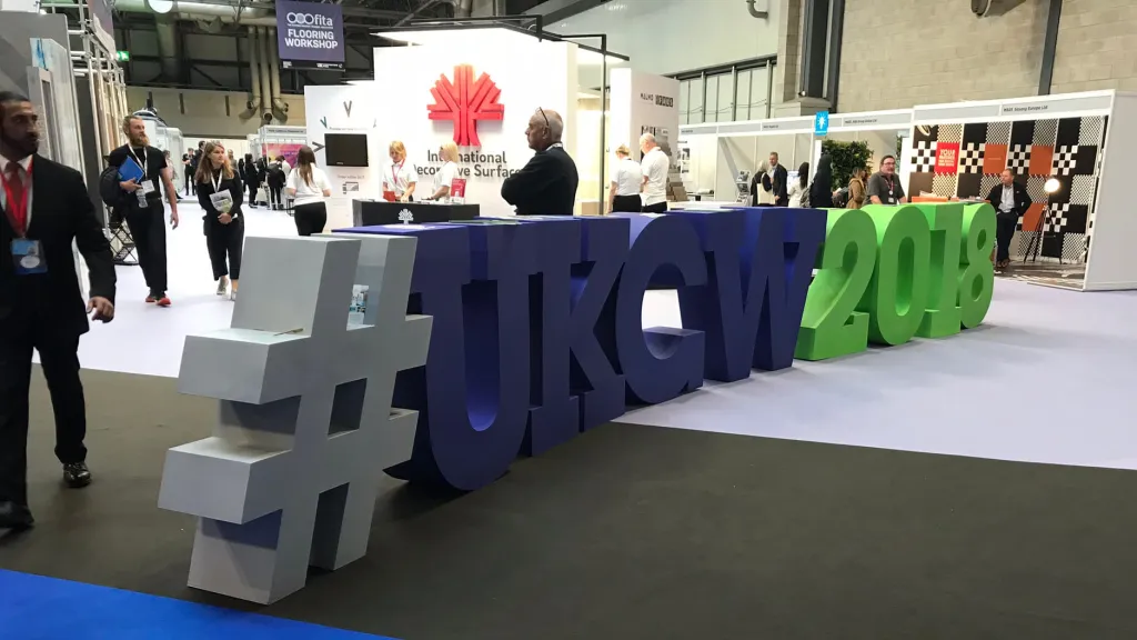 Large three-dimensional letters spelling "#UK20" in gray, navy blue, and green are displayed on a white platform at an indoor exhibition. Visitors and exhibition booths are visible in the background, with a Maple Leaf logo visible on signage behind the installation.
